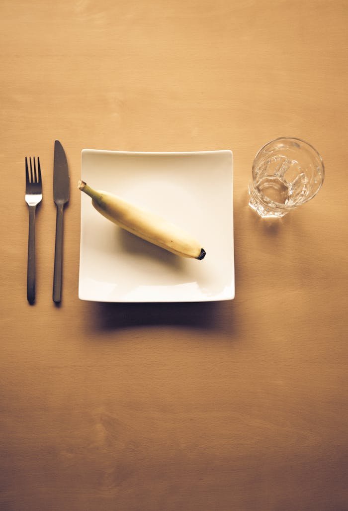 menu-09 A minimalist dining arrangement featuring a banana on a square plate, with cutlery and a glass.